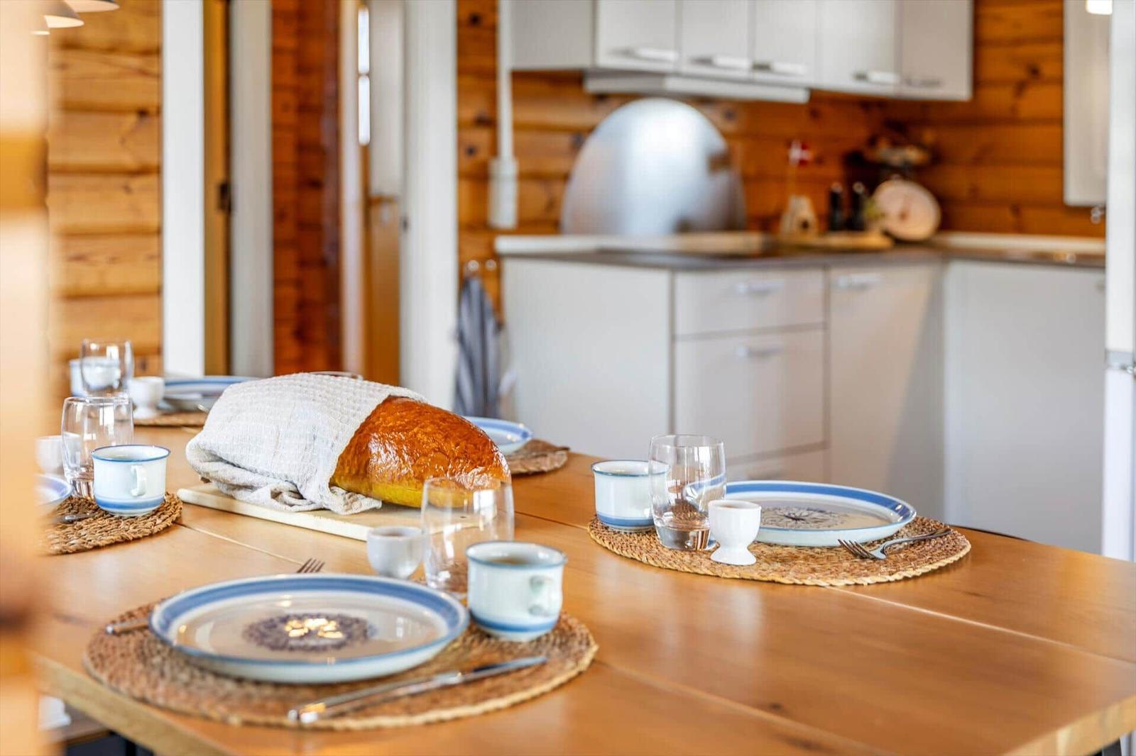 Esszimmer mit Holztisch, Tischdecken und Brot. Küche im Hintergrund.