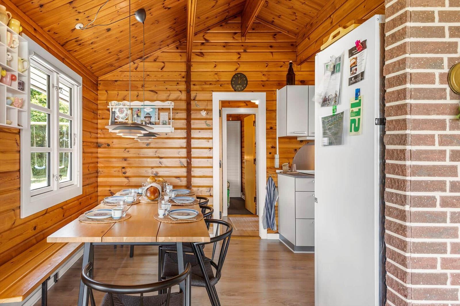 Essbereich mit Tisch, Stühlen und Küchenzeile in Holzhaus. Fenster, Kühlschrank und Wandkacheln sichtbar.