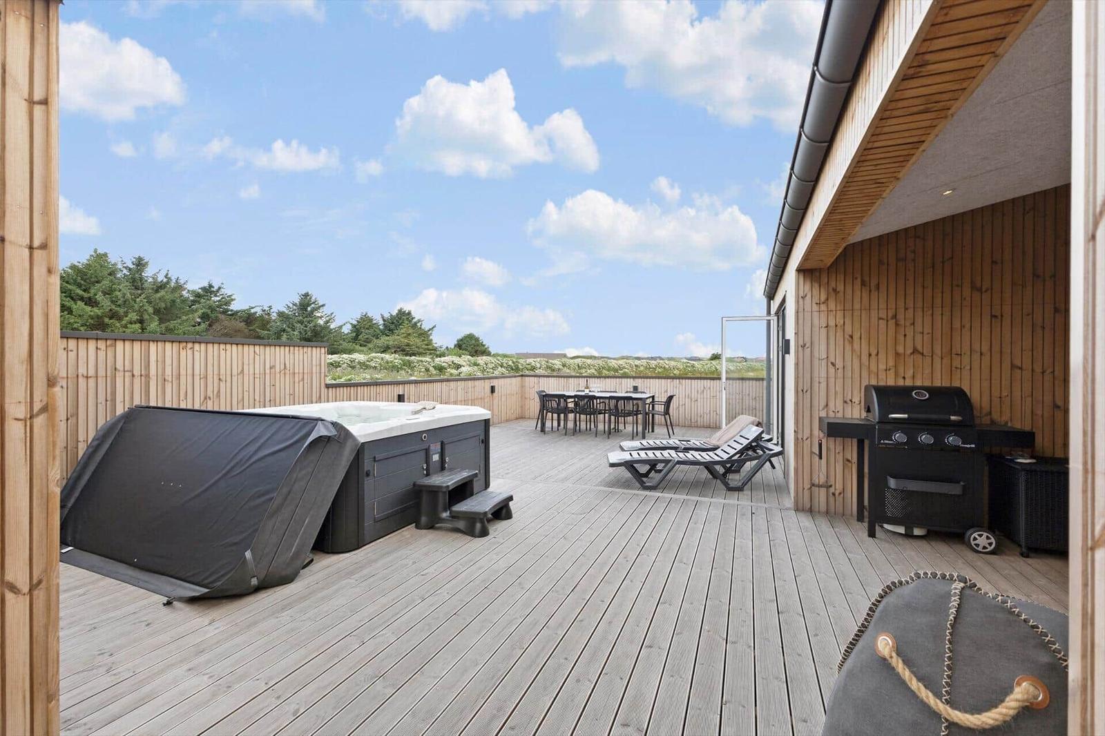 Terrasse mit Whirlpool, Liegen und Grillplatz. Holzverkleidung und Sicht auf Grünflächen.
