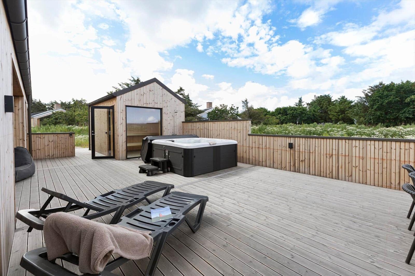 Terrasse mit Whirlpool, Liegestühlen und Holzsauna. Holzgeländer und grüne Landschaft im Hintergrund.