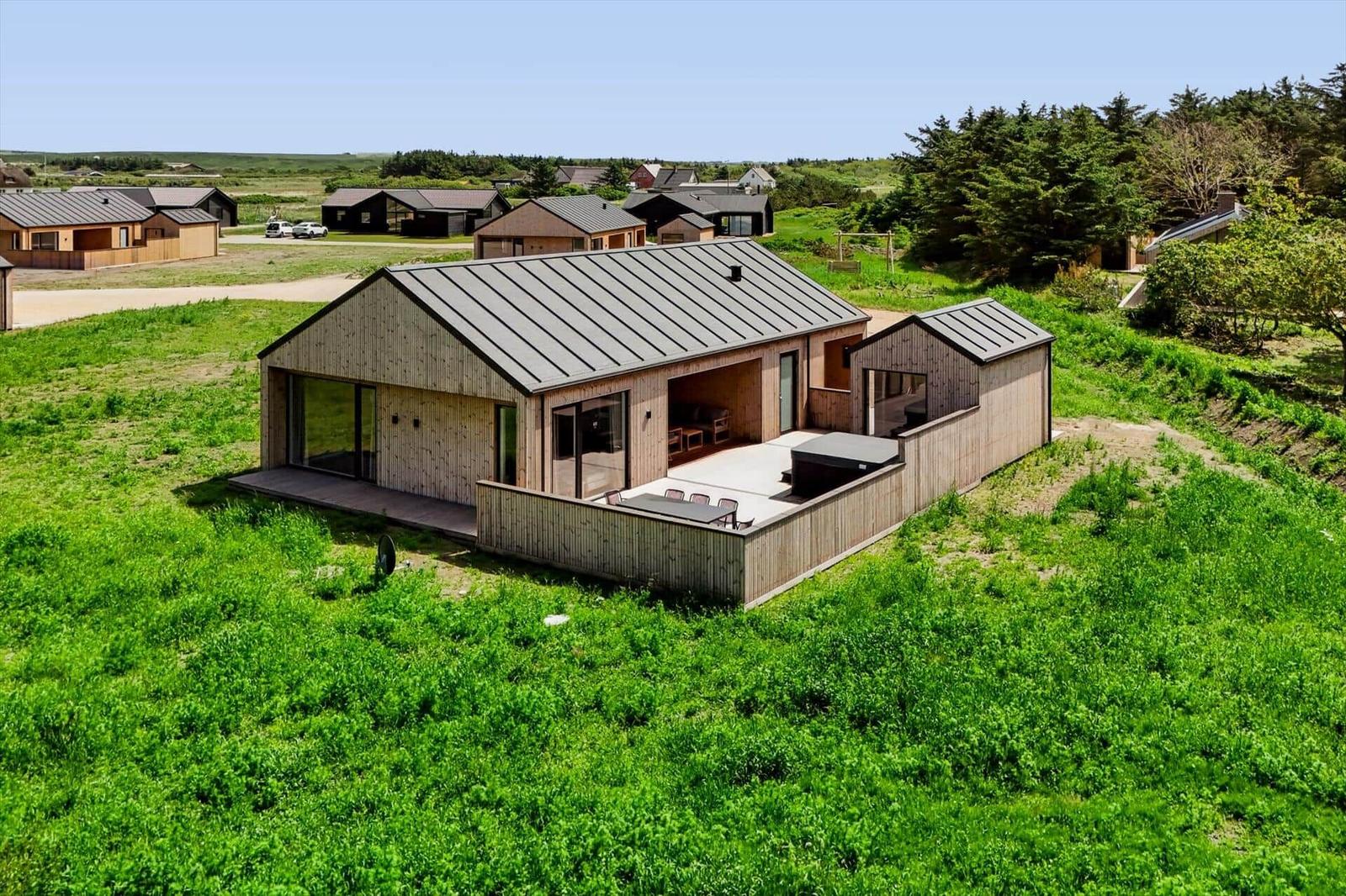 Moderne Holzhütte mit Terrasse und Garten. Im Hintergrund sind weitere Häuser und Bäume sichtbar.