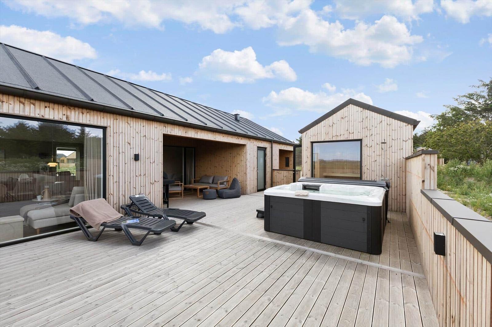 Terrasse mit Liegestühlen und Whirlpool neben Holzhaus unter blauem Himmel.