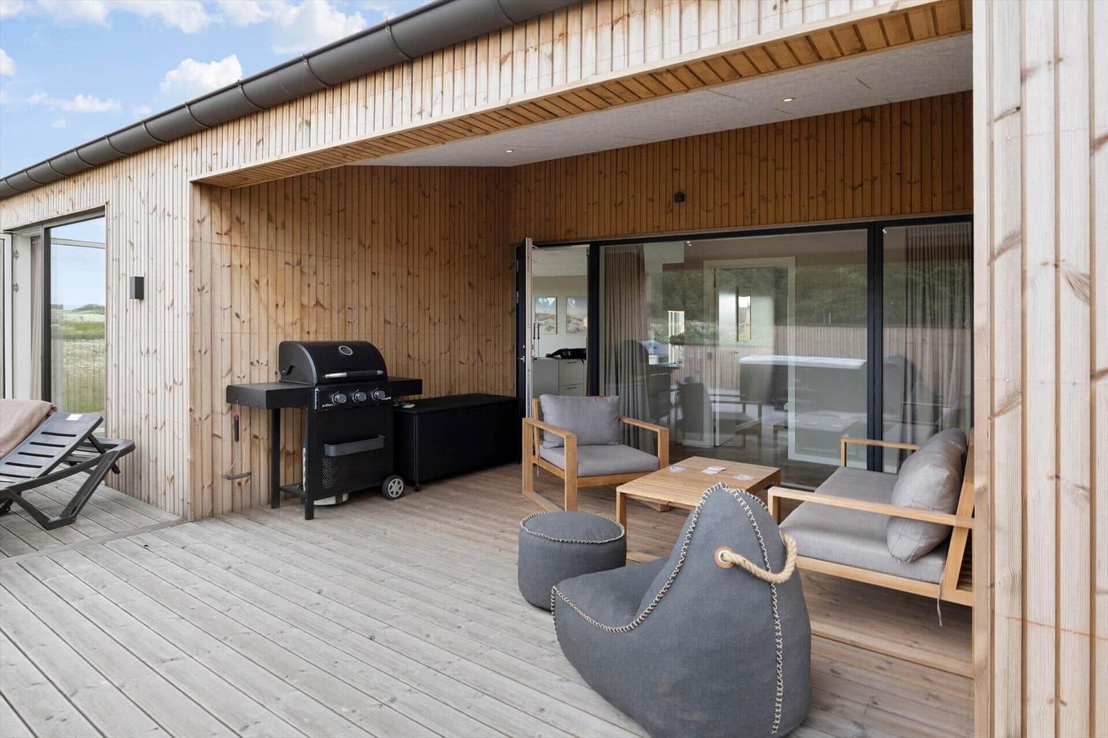 Terrasse mit Grill, Sitzgelegenheiten und Holzwand. Blick in das Innere durch Glastüren.