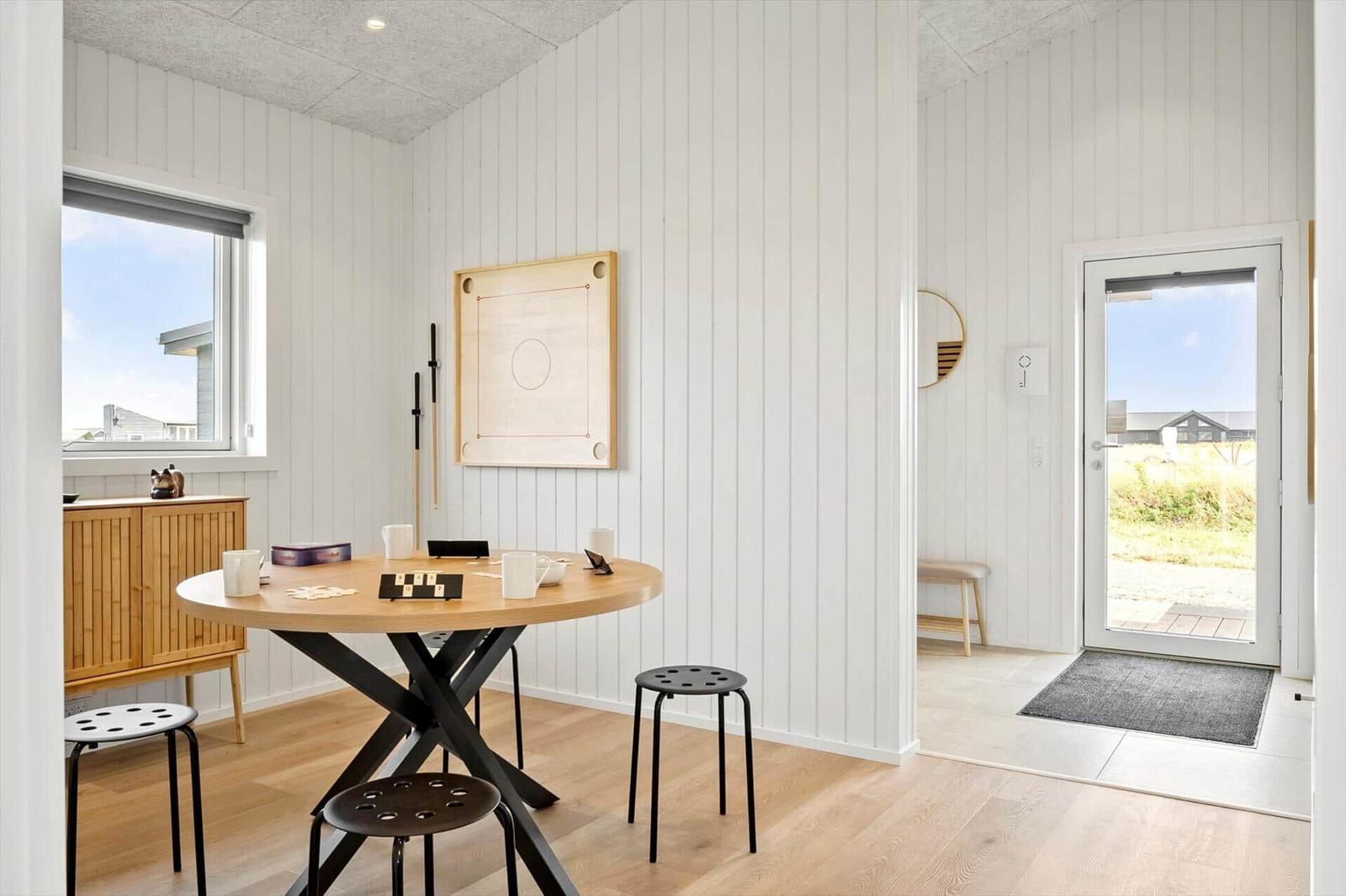 Raum mit Tisch, Stühlen und Spielplatte. Tür führt nach draußen. Fenster zeigt Außenansicht.