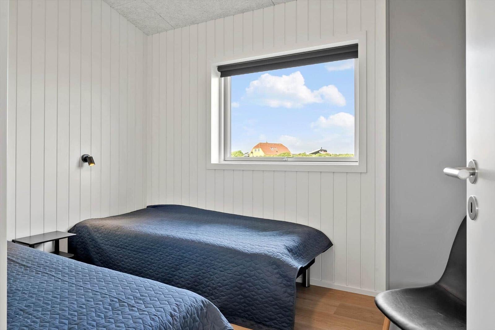 Zwei Betten mit blauen Bettdecken, Fenster mit Blick auf Haus und Himmel, Holzboden, Wandlampe und Stuhl.