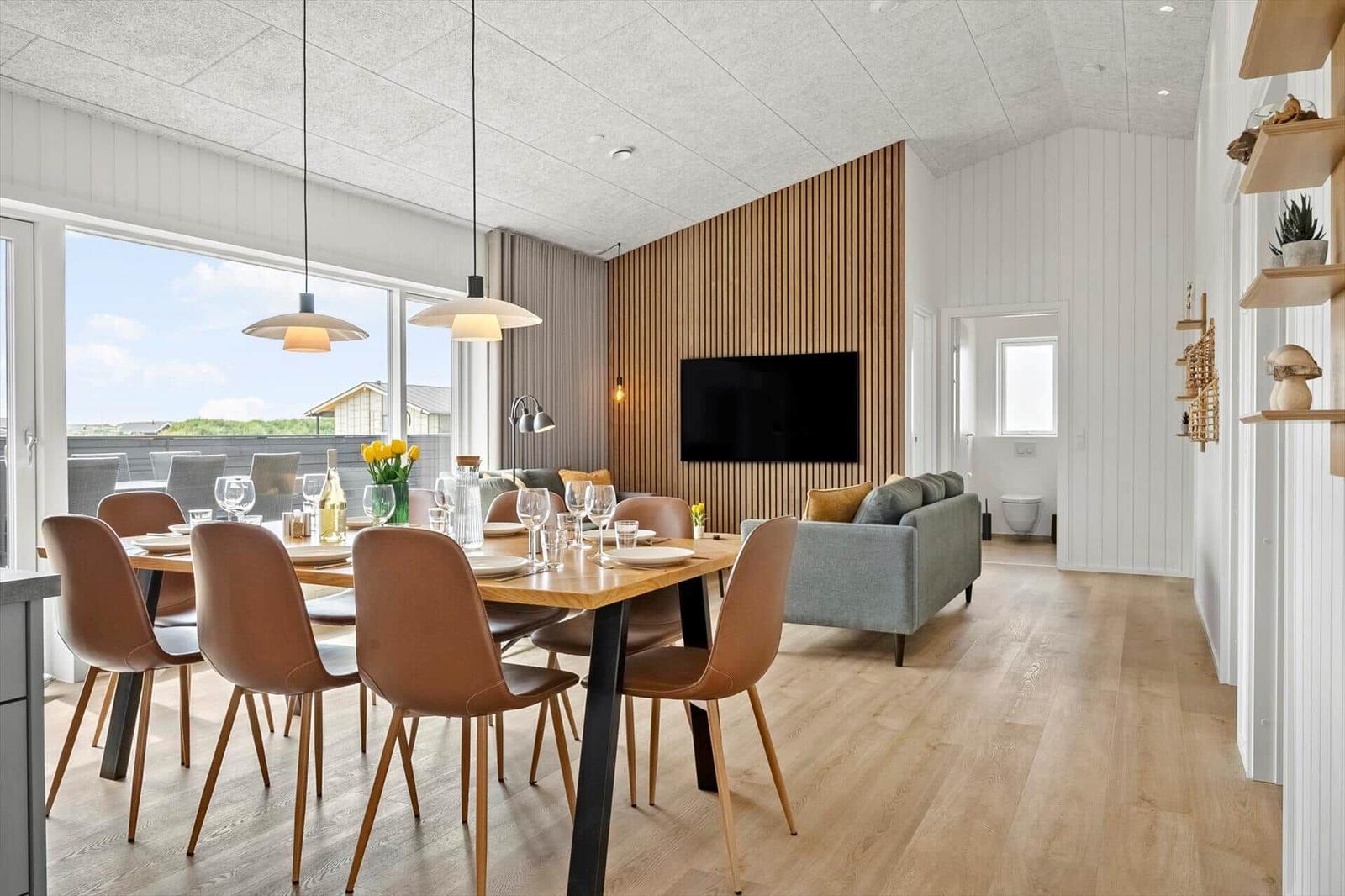 Essbereich mit Tisch und Stühlen, Wohnbereich mit Sofa und Fernseher, Holzfußboden und große Fenster.