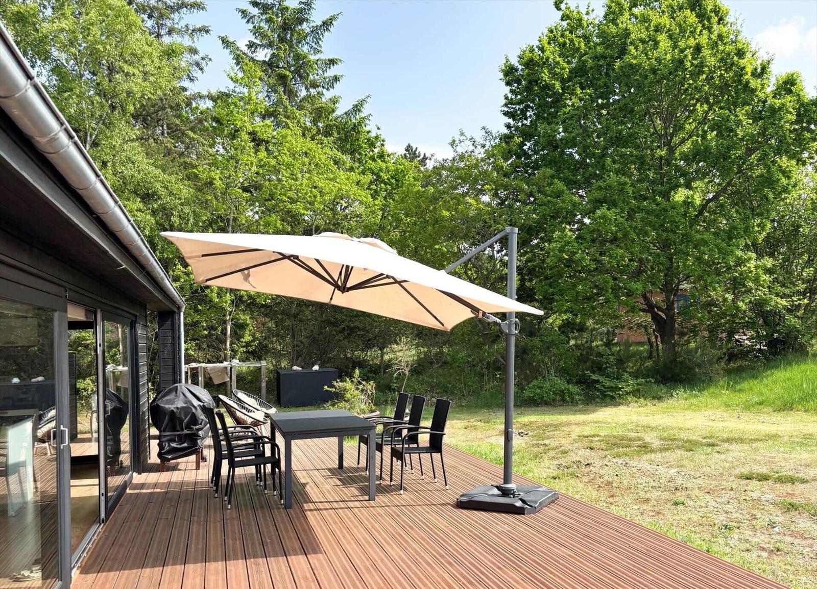 Terrasse mit Tisch, Stühlen und Sonnenschirm. Hintergrund: grüne Bäume und Rasen.