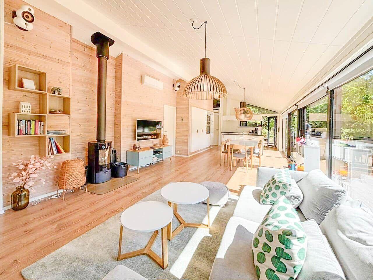 Wohnbereich mit Holzwand, Kamin, Sofa und Essbereich. Große Fenster mit Blick auf Garten.