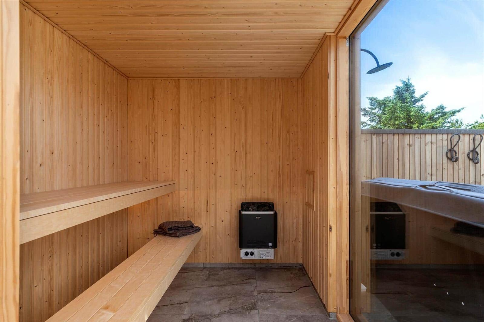 Sauna mit Holzwand, Bank und Heizgerät. Durchgang zu Außenbereich mit Dusche.