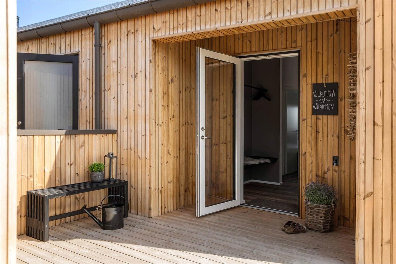 Holzveranda mit Glastür und Willkommensschild. Bank, Topf und Gießkanne stehen bereit.