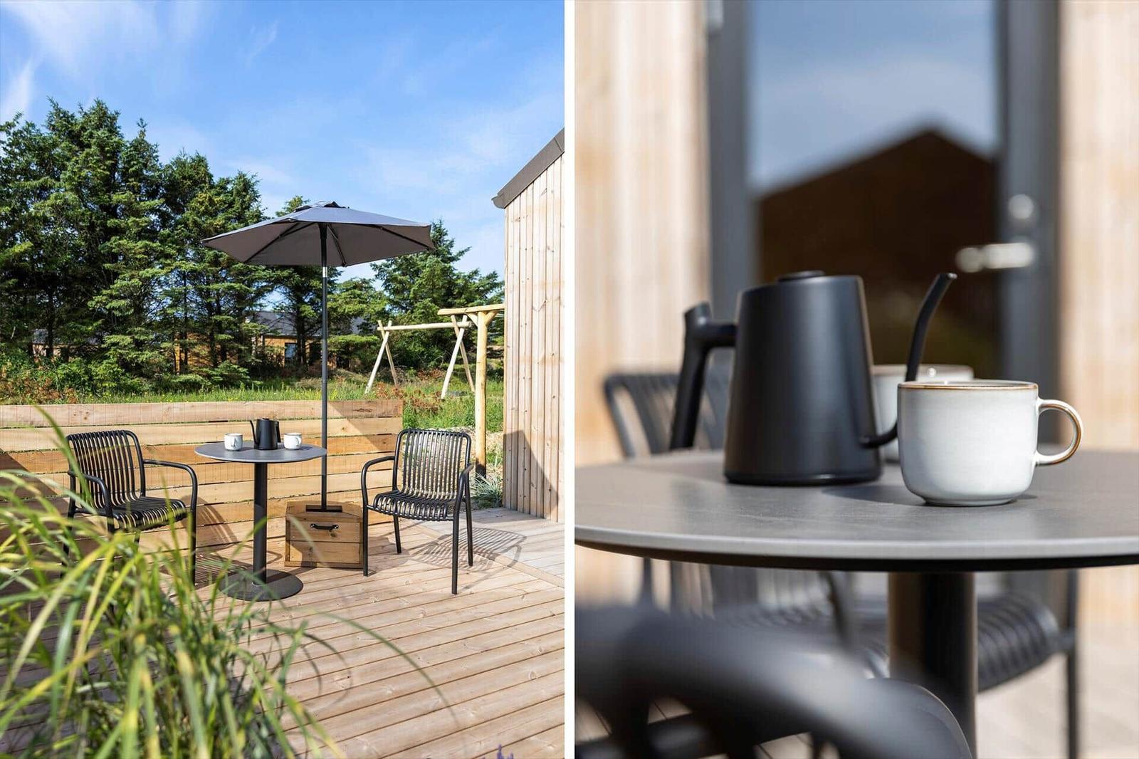 Terrasse mit Tisch, zwei Stühlen und Sonnenschutz. Im Hintergrund sind Bäume und ein Schaukelgerüst sichtbar.