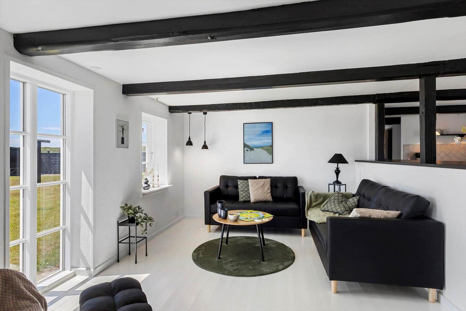 Wohnzimmer mit schwarzen Sofas, Holzbalken und großem Fenster. Runder Teppich und Tisch.