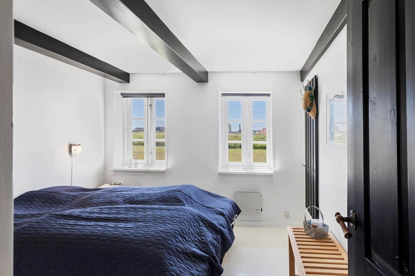 Schlafzimmer mit blauem Bett, zwei Fenstern und schwarzem Balken an der Decke.
