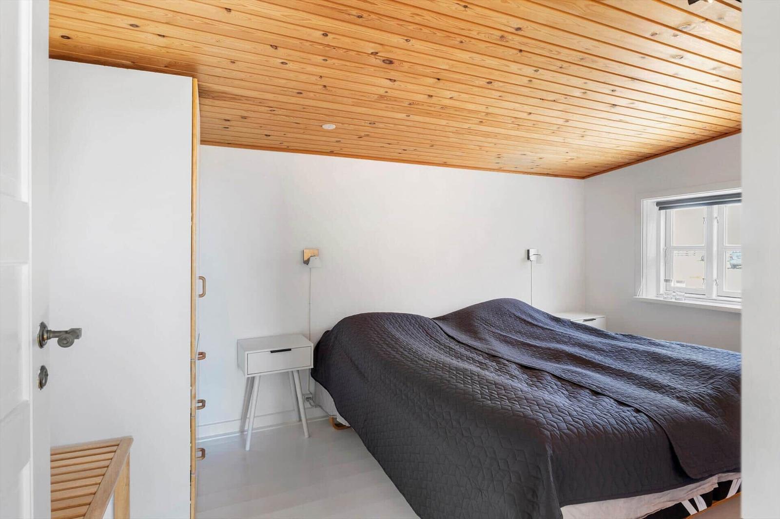 Schlafzimmer mit Holzdecke, Bett mit dunkler Bettwäsche und kleinem Nachttisch.