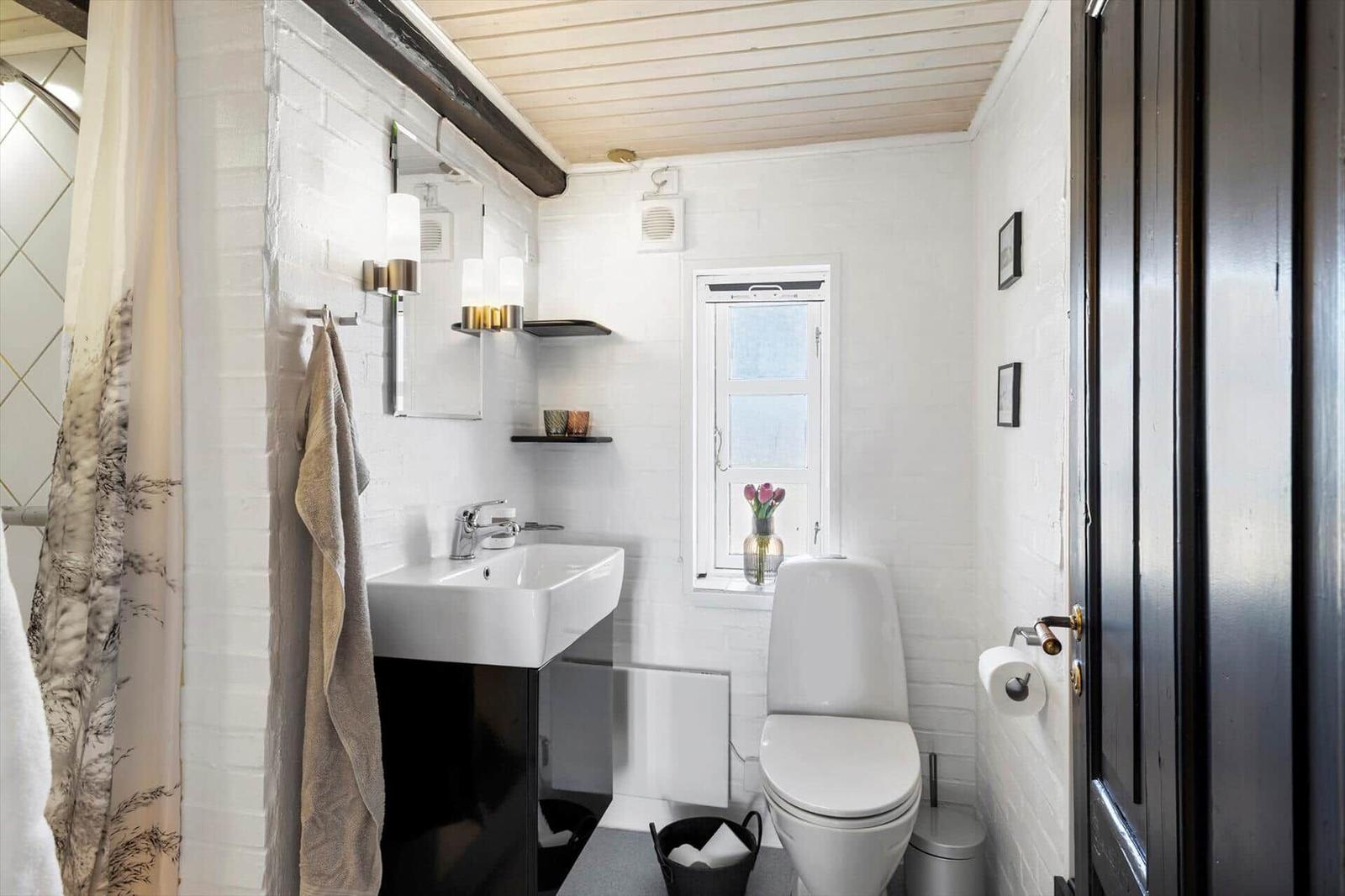 Badezimmer mit Toilette, Waschbecken und Fenster. Weiße Wände und Holzdecke.