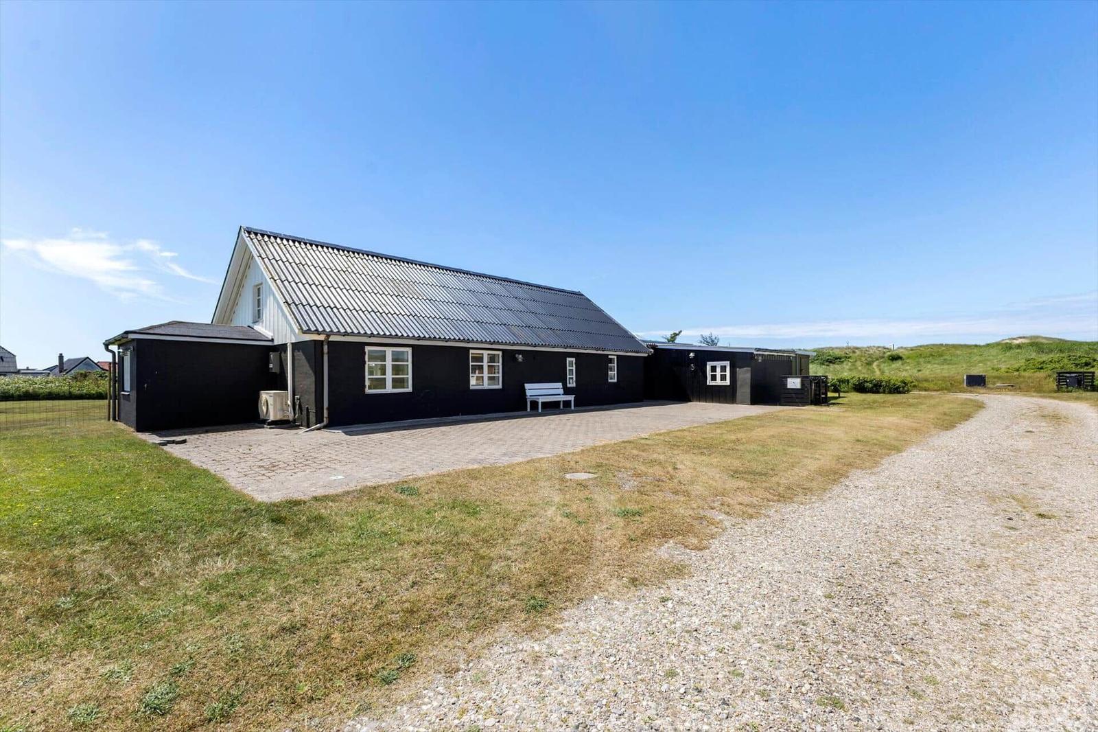 Schwarzes Ferienhaus mit Dachrinne, weißer Bank und gepflastertem Hof. Grasfläche und Kiesweg.