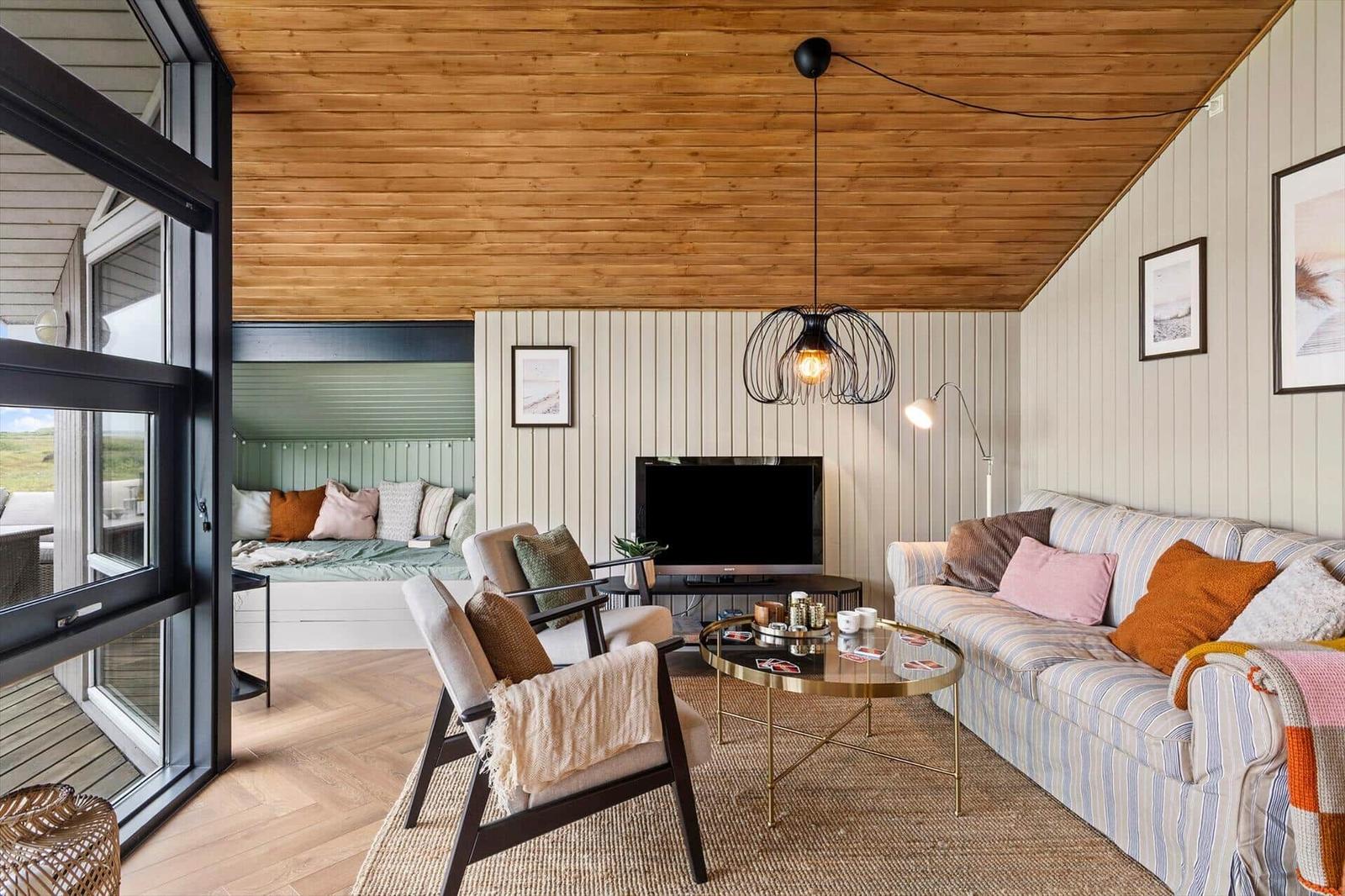 Wohnzimmer mit Sofa, Sessel und Fernseher. Holzdecke und großes Fenster.