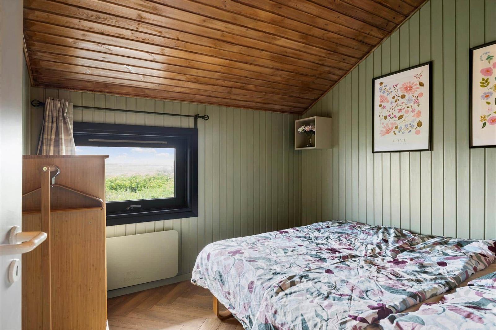 Schlafzimmer mit Holzdecke, grünen Wänden, Bett mit Blumenbezug und Fenster mit Blick auf Landschaft.