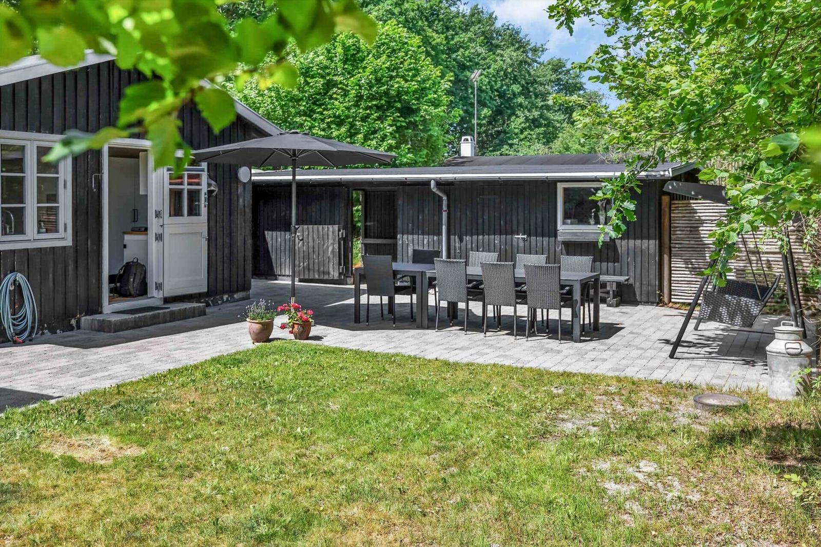 Haus mit Terrasse, Tisch, Stühlen und Sonnenschirm. Grasfläche und Bäume im Hintergrund.