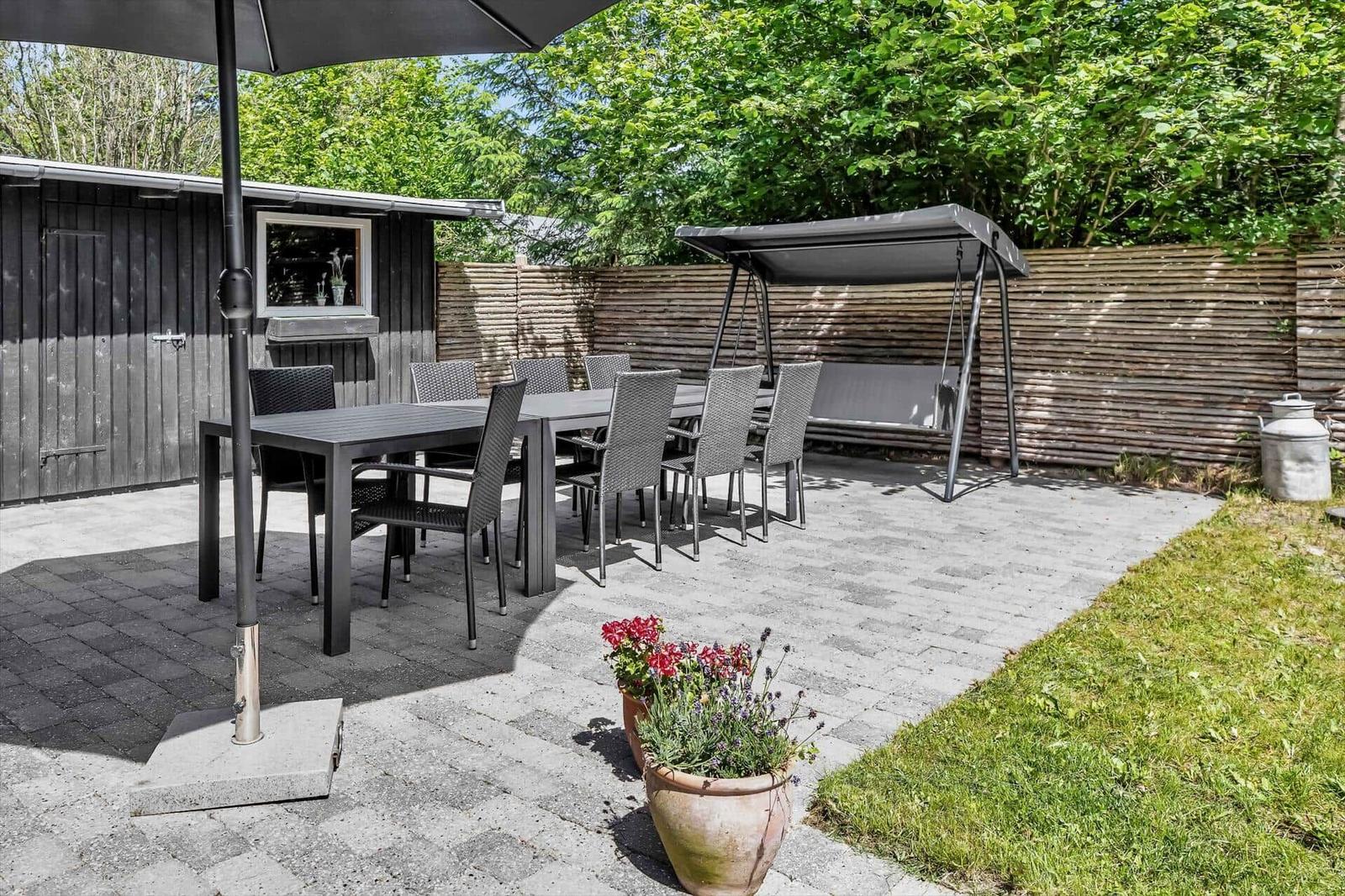 Terrasse mit Tisch, Stühlen, Sonnenschirm und Schaukel unter Bäumen.