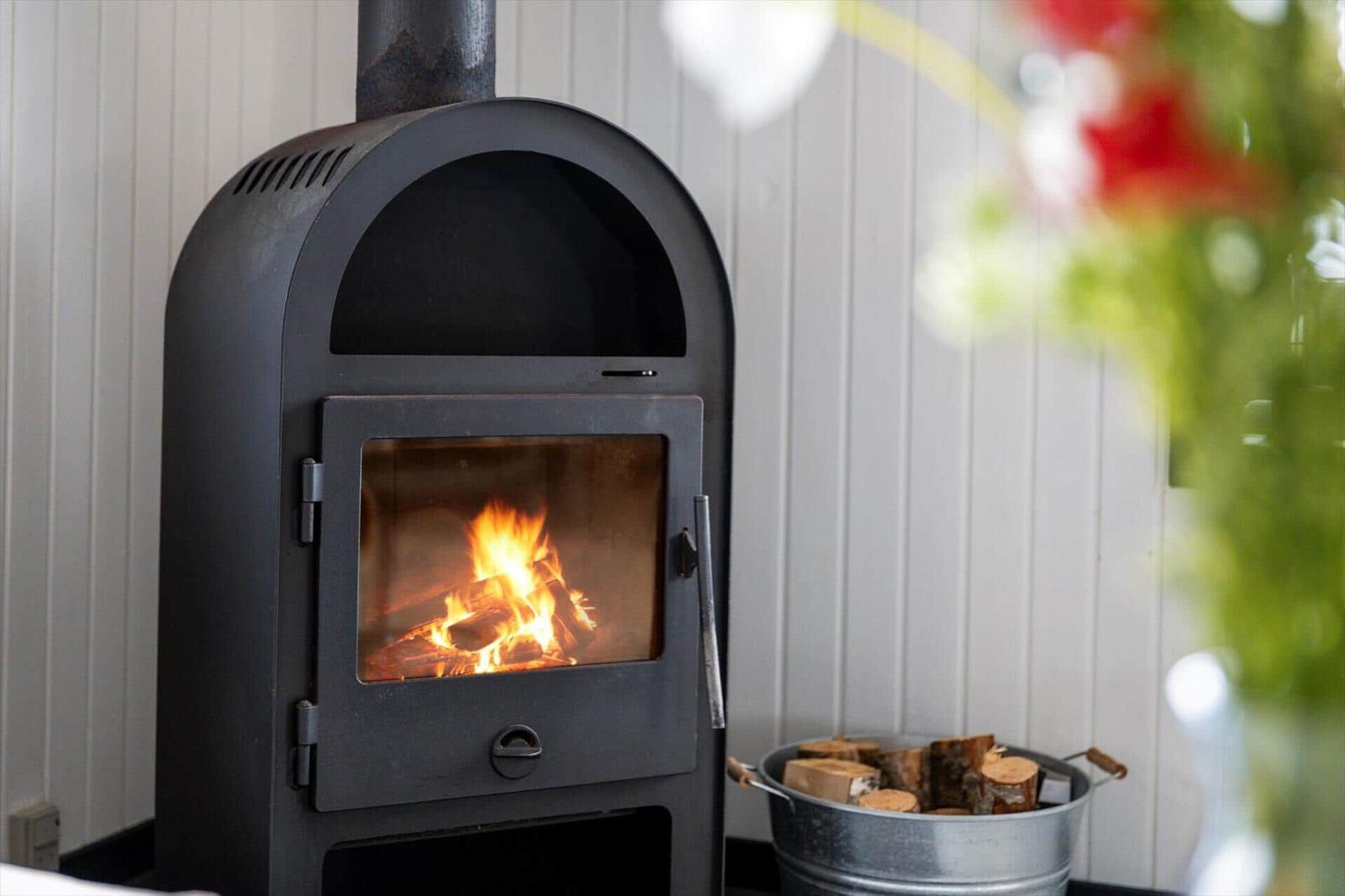 Ein schwarzer Holzofen mit brennendem Feuer steht vor einer weißen Wand. Daneben steht ein Eimer mit Brennholz.