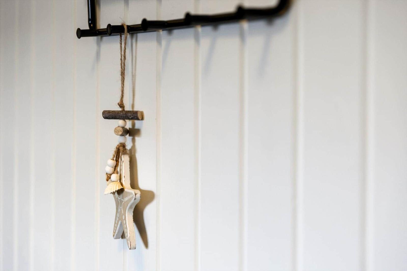 Ein Holzstern mit Muschel und Perlen hängt an einer schwarzen Stange an einer weißen Wand.