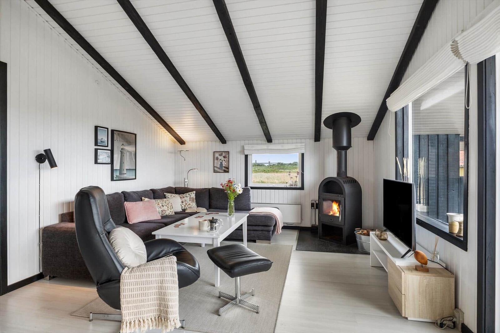 Wohnzimmer mit Sofa, Sessel, Kamin und TV. Weiße Wände, schwarze Balken und große Fenster.
