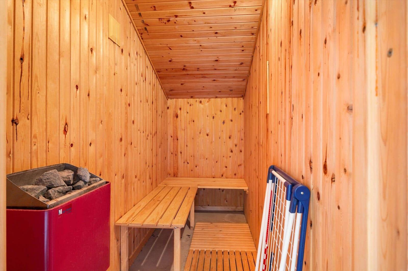 Sauna mit Holzwand, Bänken und Steinkasten. Kleiner Körbchenständer steht an der Wand.