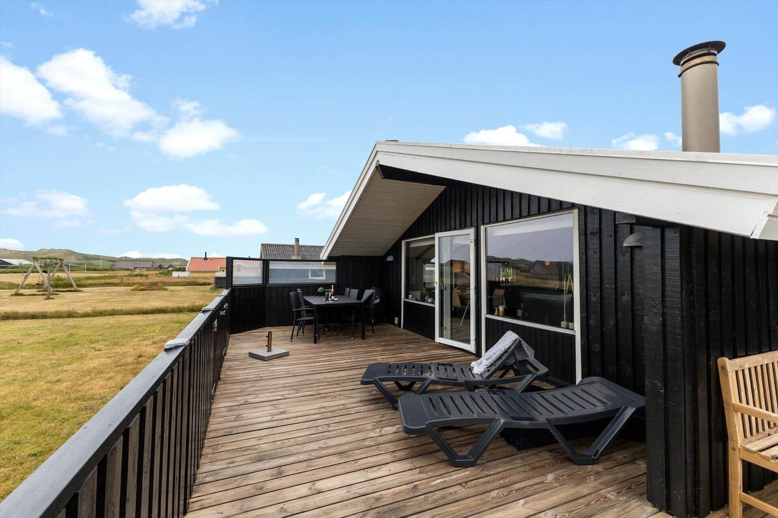 Terrasse mit Liegestühlen und Tisch. Schwarze Holzwand und weißer Dachvorsprung. Blick auf Felder.