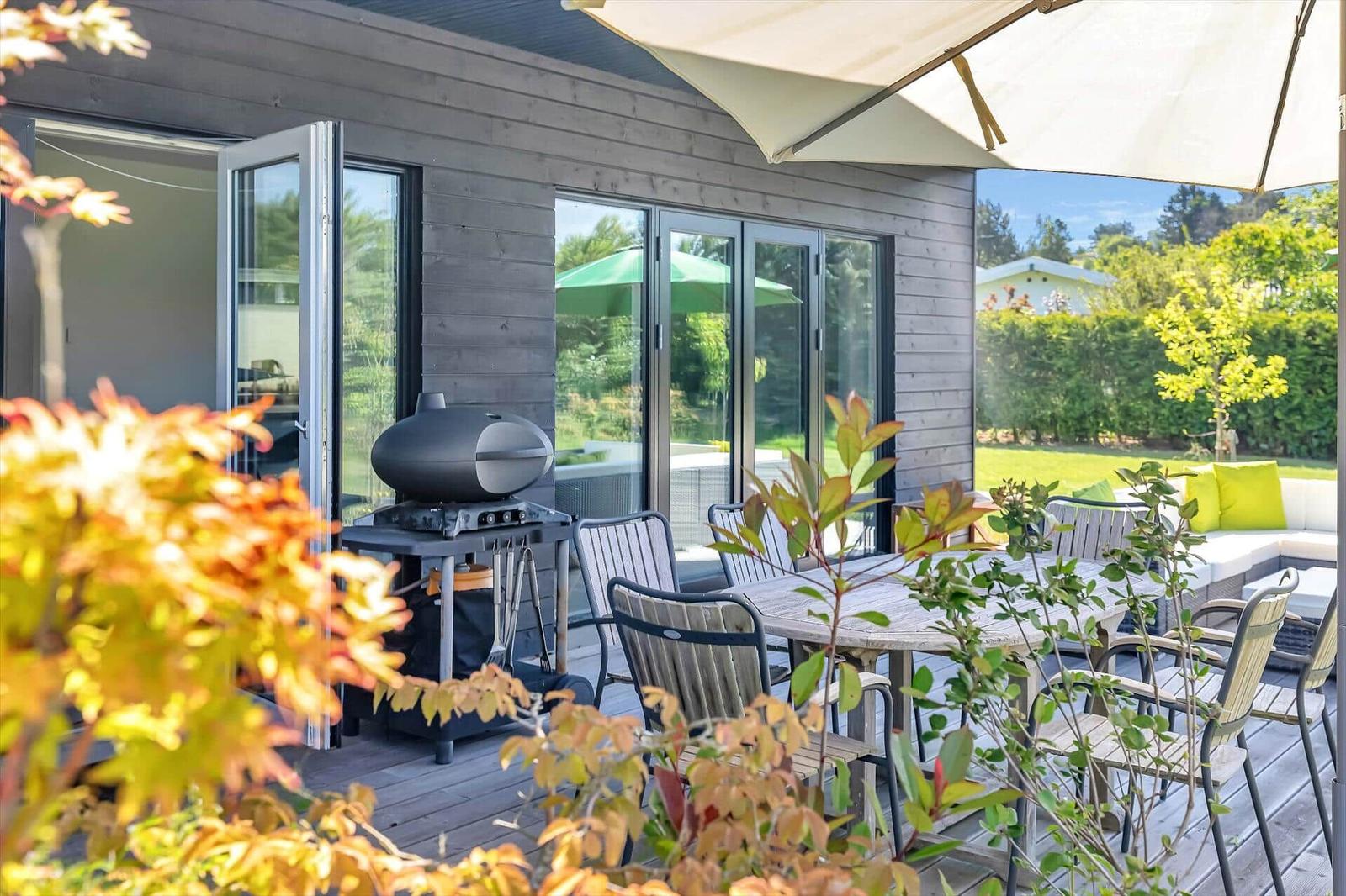 Terrace with table, chairs, umbrella, and grill. Background: garden with green areas and trees.