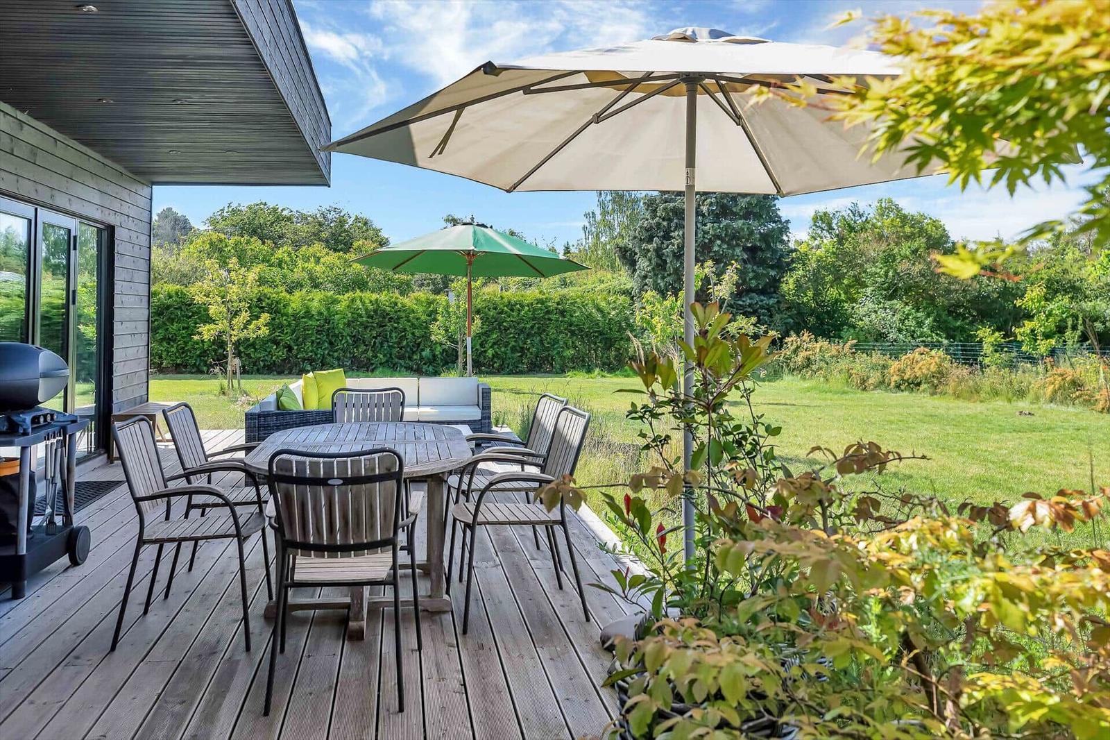 Deck with table, chairs, and umbrella. Lawn and hedges in the background.