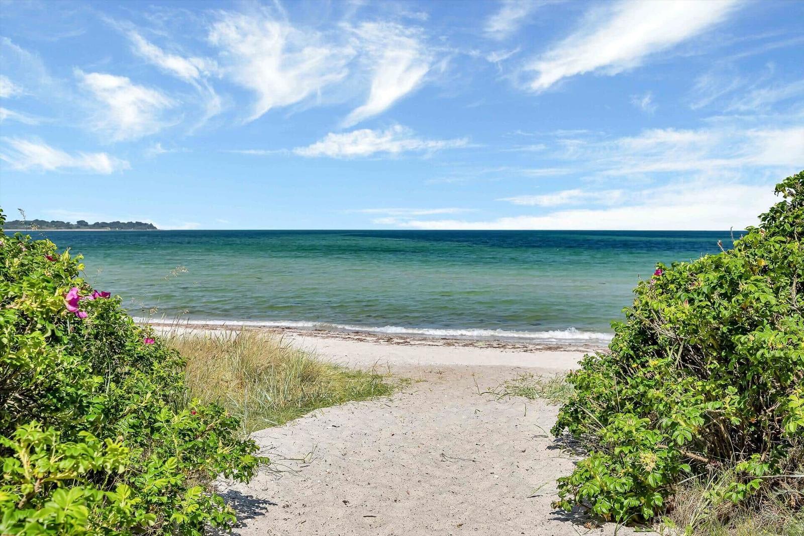 Sandy path leads to the beach with clear sea and blue sky.