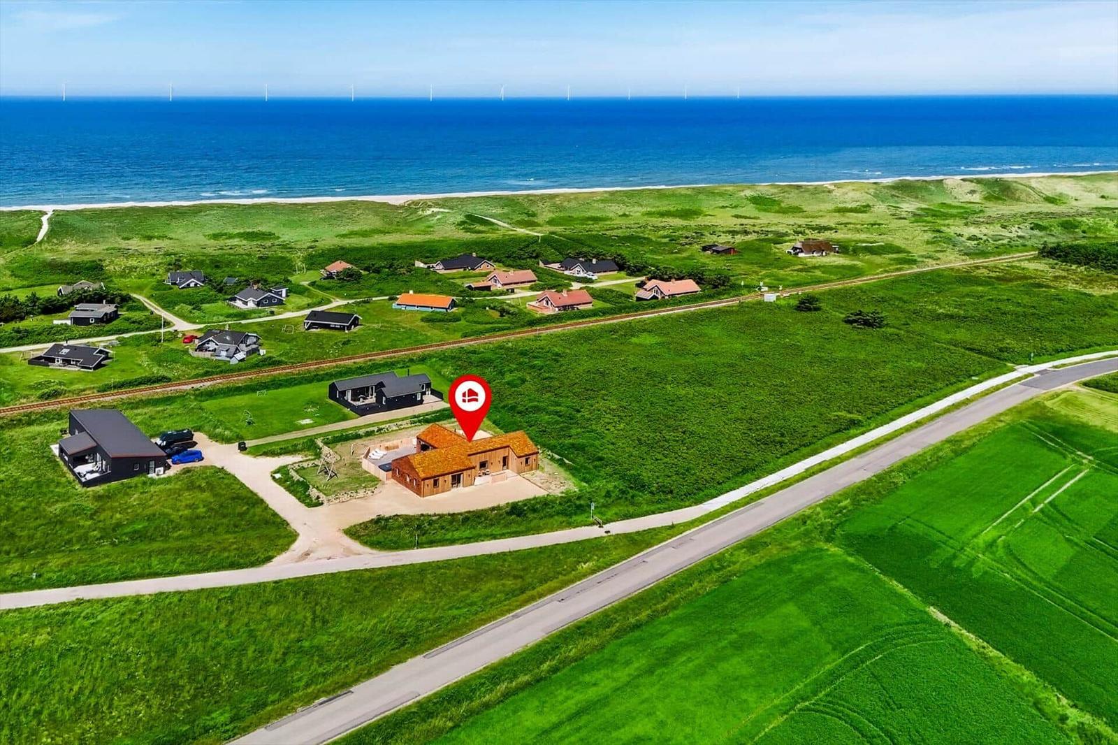 Haus mit rotem Dach liegt in grüner Landschaft neben Strand und Windkraftanlagen.