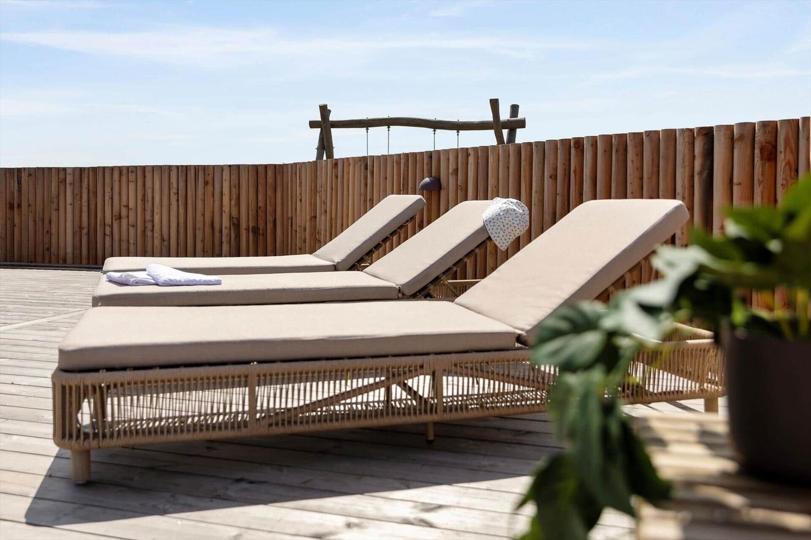 Drei Liegestühle auf einer Holzterrasse mit Holzgeländer und Pflanze im Vordergrund.