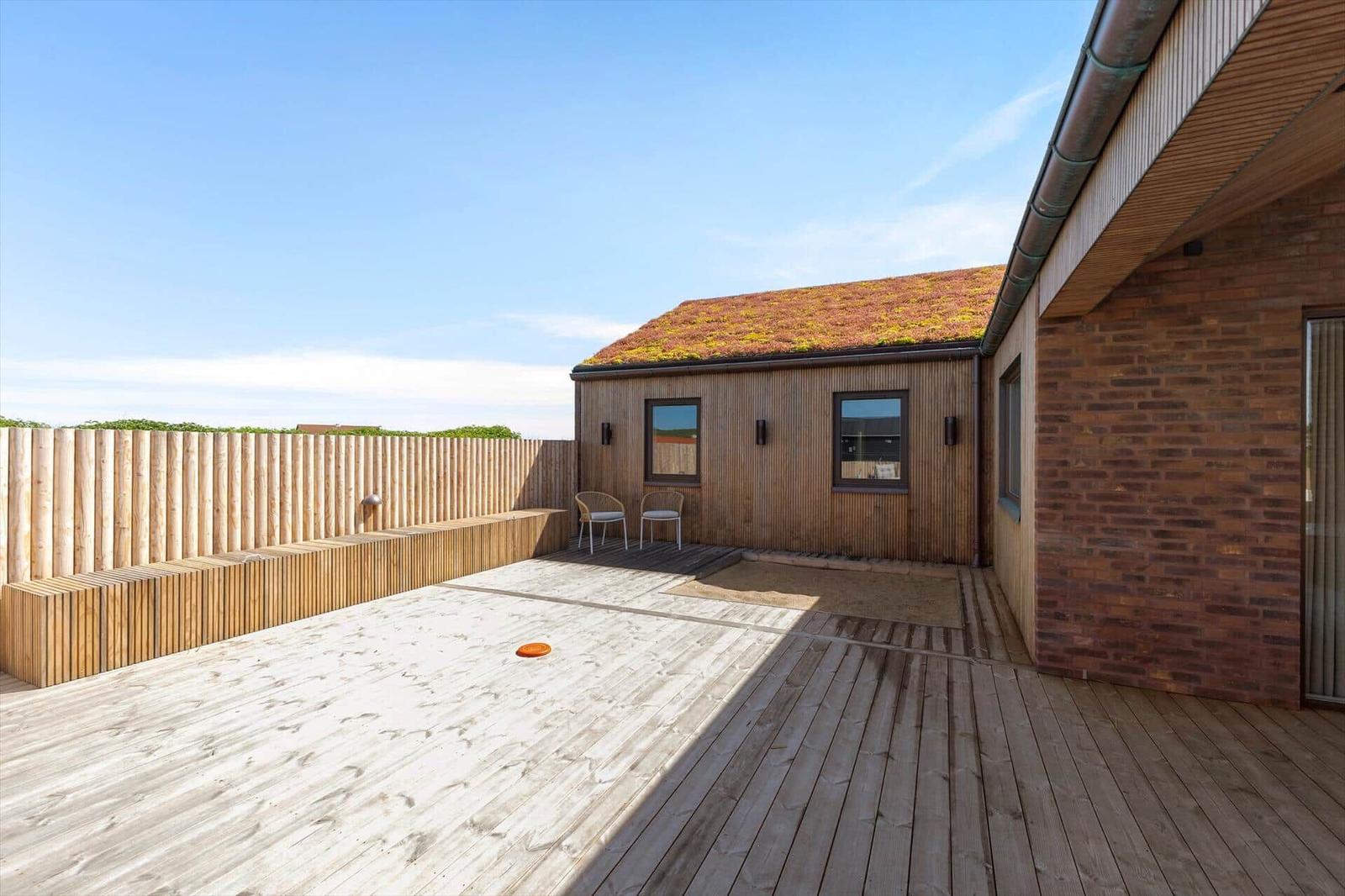 Terrasse mit Holzdecke, Sitzgelegenheit und Gartenzaun. Hintergrund: Holzhaus mit grüner Dachdeckung.