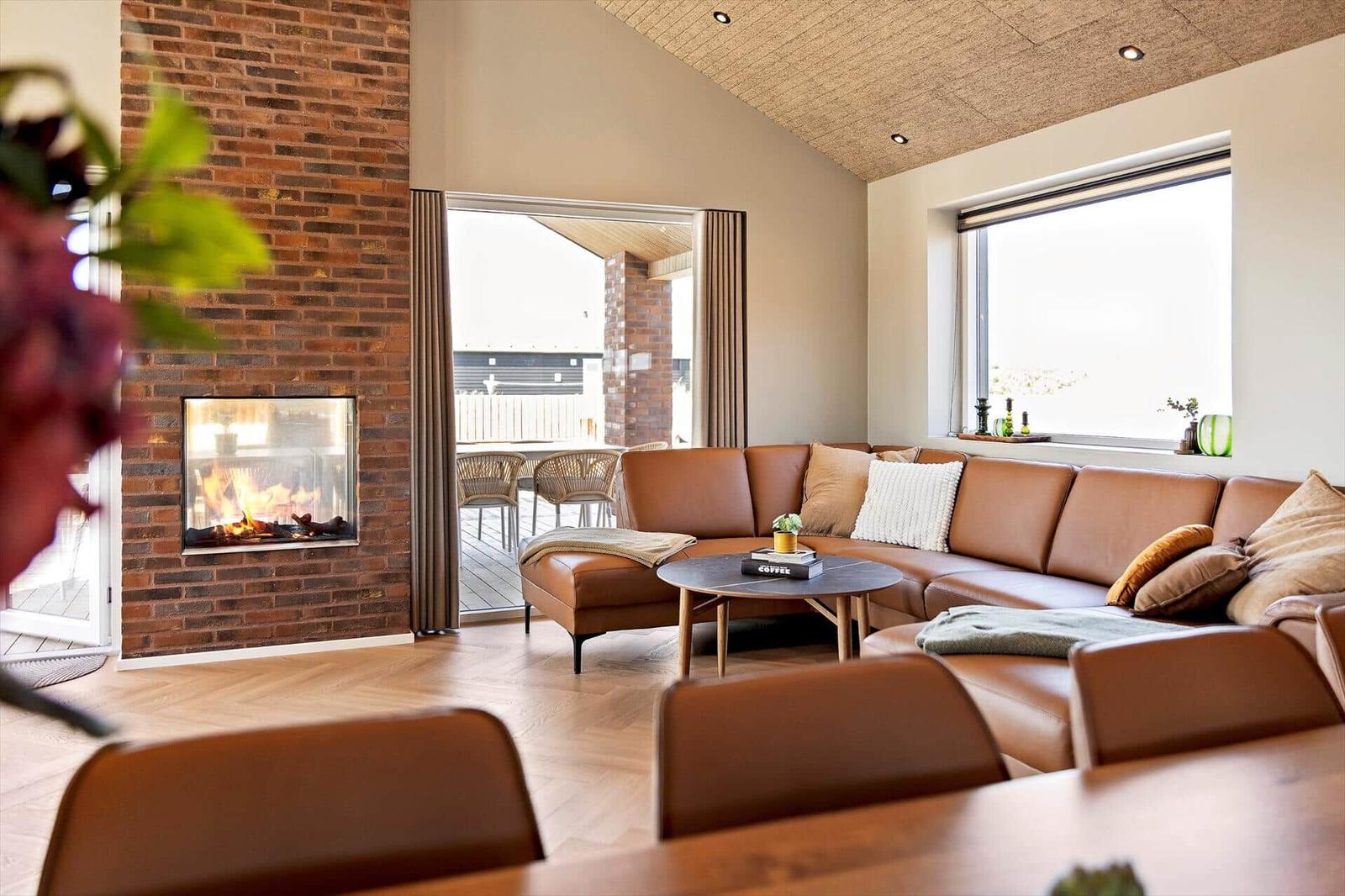 Wohnzimmer mit braunem Sofa, Kamin und großem Fenster. Holzboden und hölzerner Tisch.