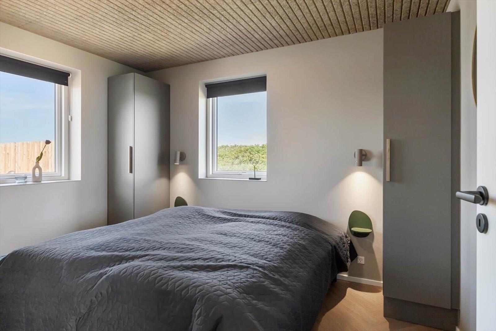 Schlafzimmer mit Bett, zwei Fenstern und zwei Schränken. Decke aus Holzlamellen.