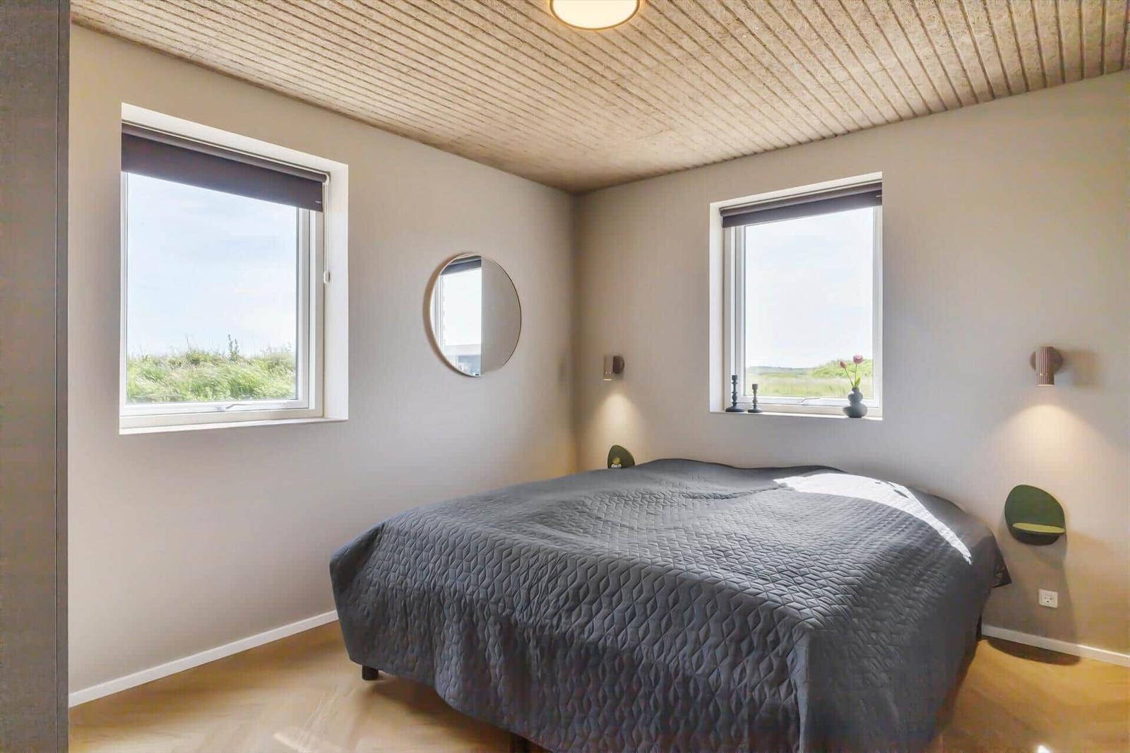 Schlafzimmer mit Doppelbett, zwei Fenstern und runden Spiegel an den Wänden.