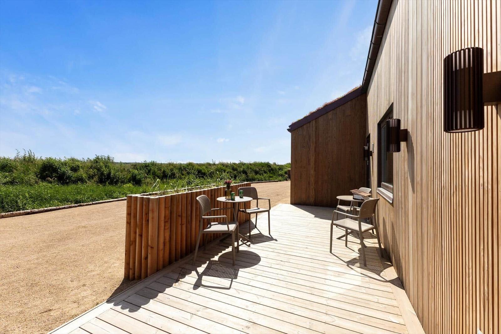 Terrasse mit Tischen und Stühlen neben Holzhaus. Blick auf Grünfläche und Himmel.