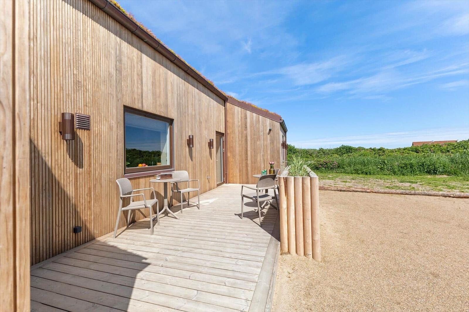 Holzhaus mit Terrasse, Tisch und Stühlen. Grünflächen im Hintergrund.