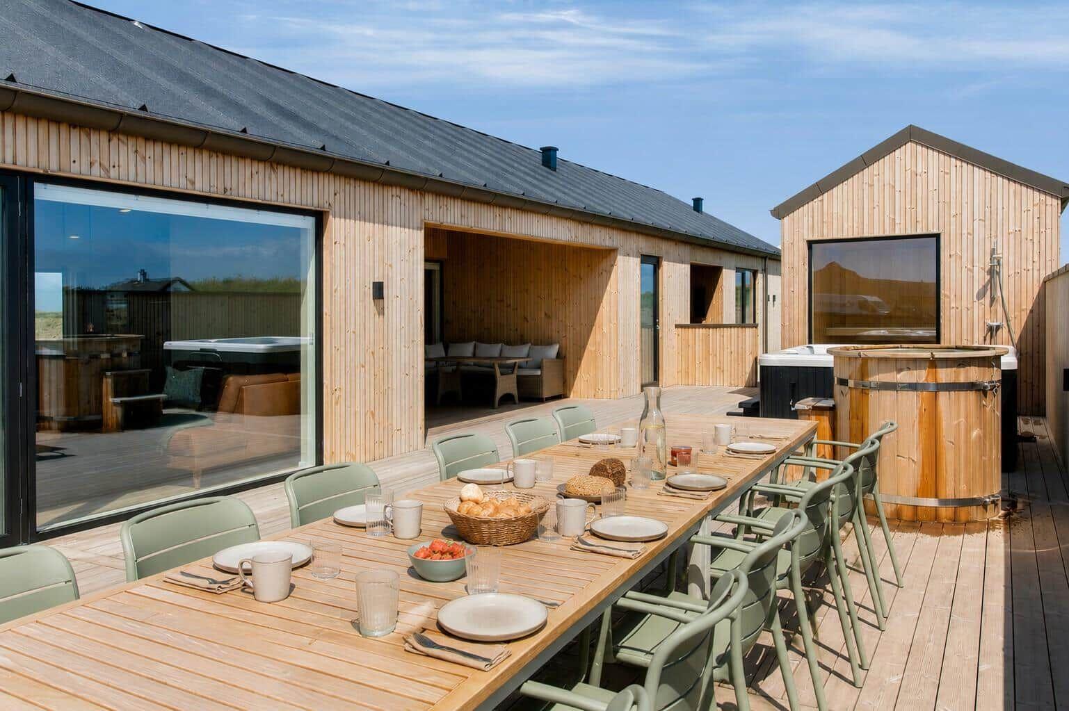 Lange Holztisch mit Stühlen auf Terrasse. Hintergrund: Holzhäuser mit Fenstern und Whirlpool.