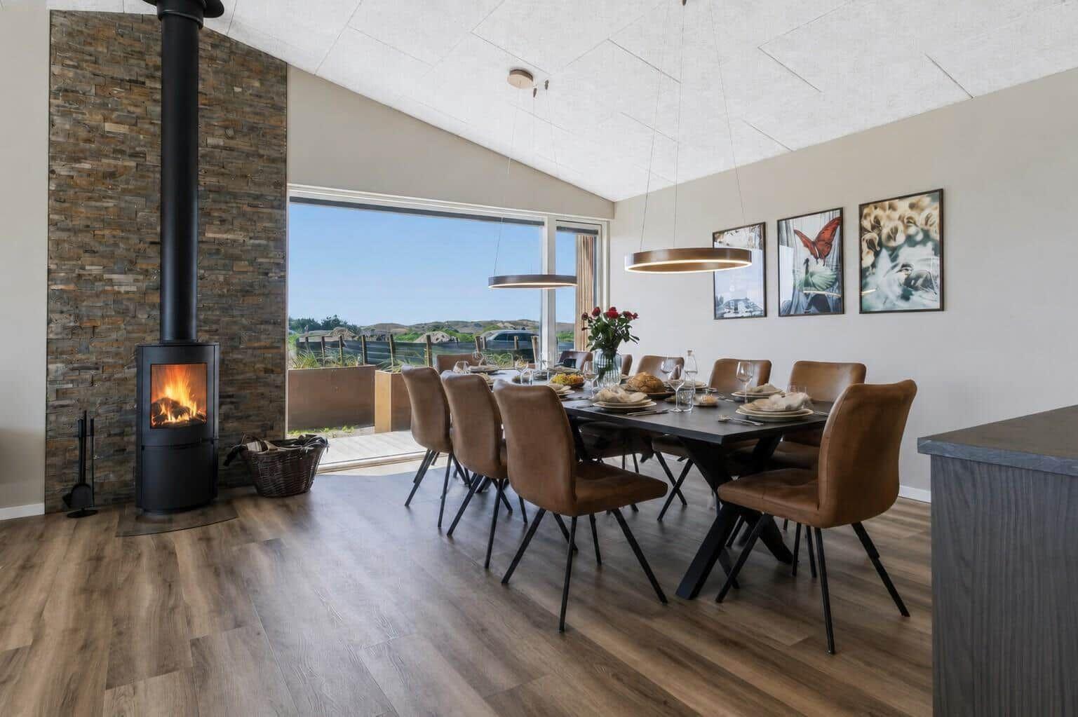 Esszimmer mit Holzboden, Esstisch, Stühlen und Kaminofen. Große Fenster bieten Blick auf Landschaft.