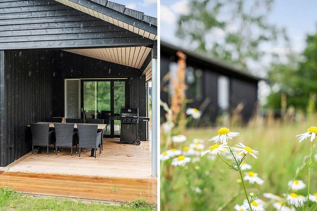 Terrasse mit Tisch, Stühlen und Grill unter schwarzem Dach. Im Hintergrund Blumenfeld und Holzhaus.