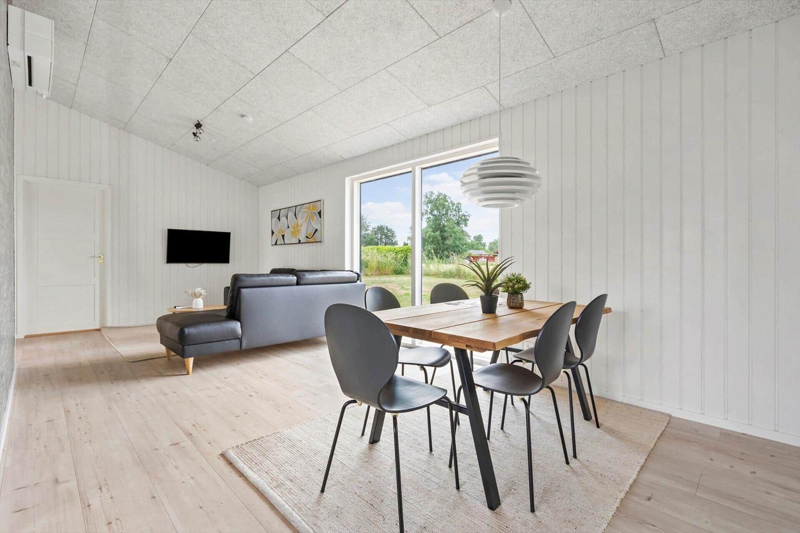 Wohn- und Essbereich mit Sofa, Esstisch und großen Fenstern. Holzboden und weiße Wände.