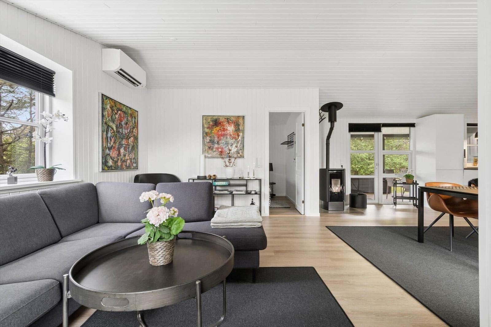 Wohnzimmer mit grauem Sofa, Holzboden und Kamin. Fenster zeigen grüne Bäume.