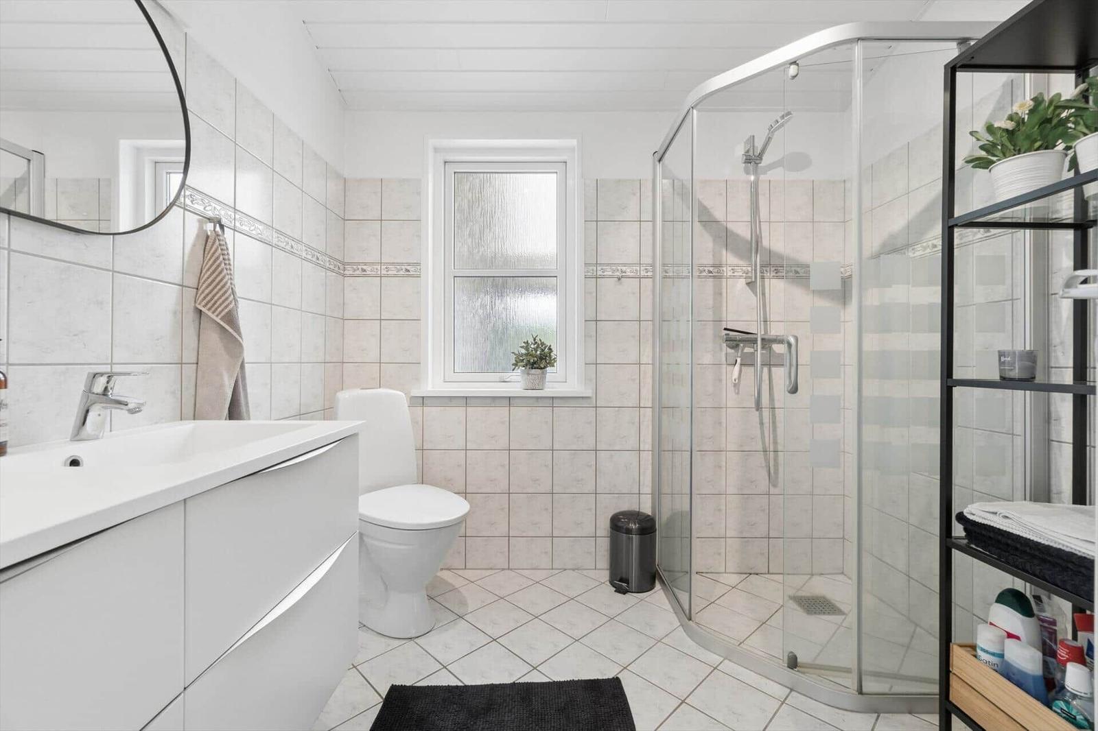 Badezimmer mit Dusche, Toilette und Waschbecken. Fenster und Regal sind sichtbar.