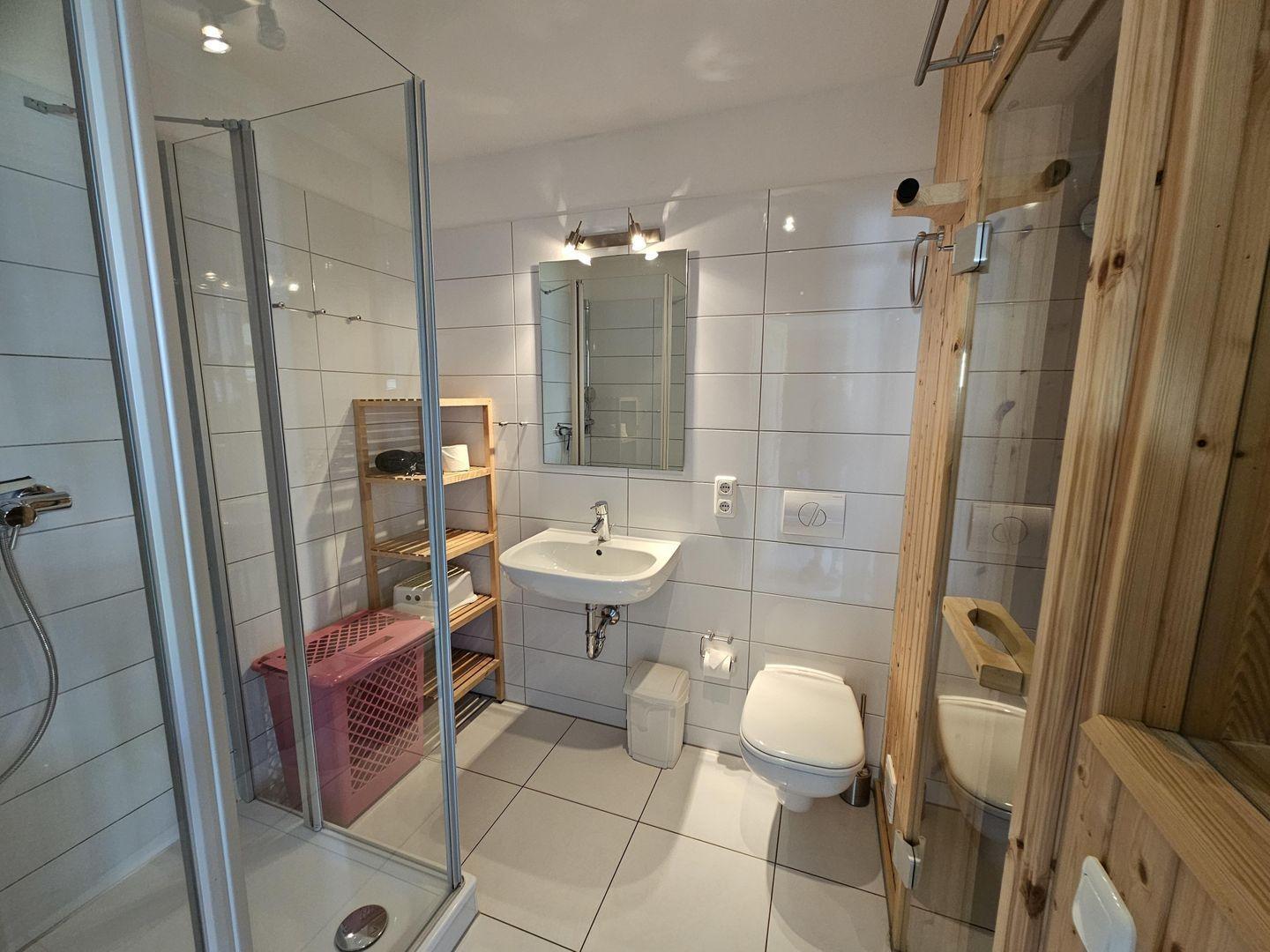 Badezimmer mit Dusche, Waschbecken und Toilette. Holzregal und rosa Wäschekorb.
