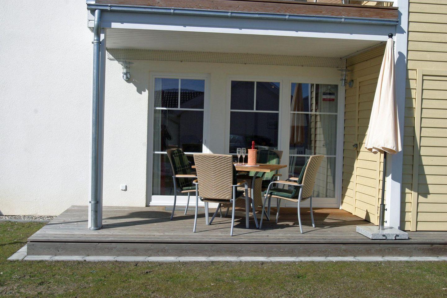 Terrasse mit Tisch, Stühlen und geschlossenem Sonnenschirm. Hintergrund: Fenster und Hauswand.