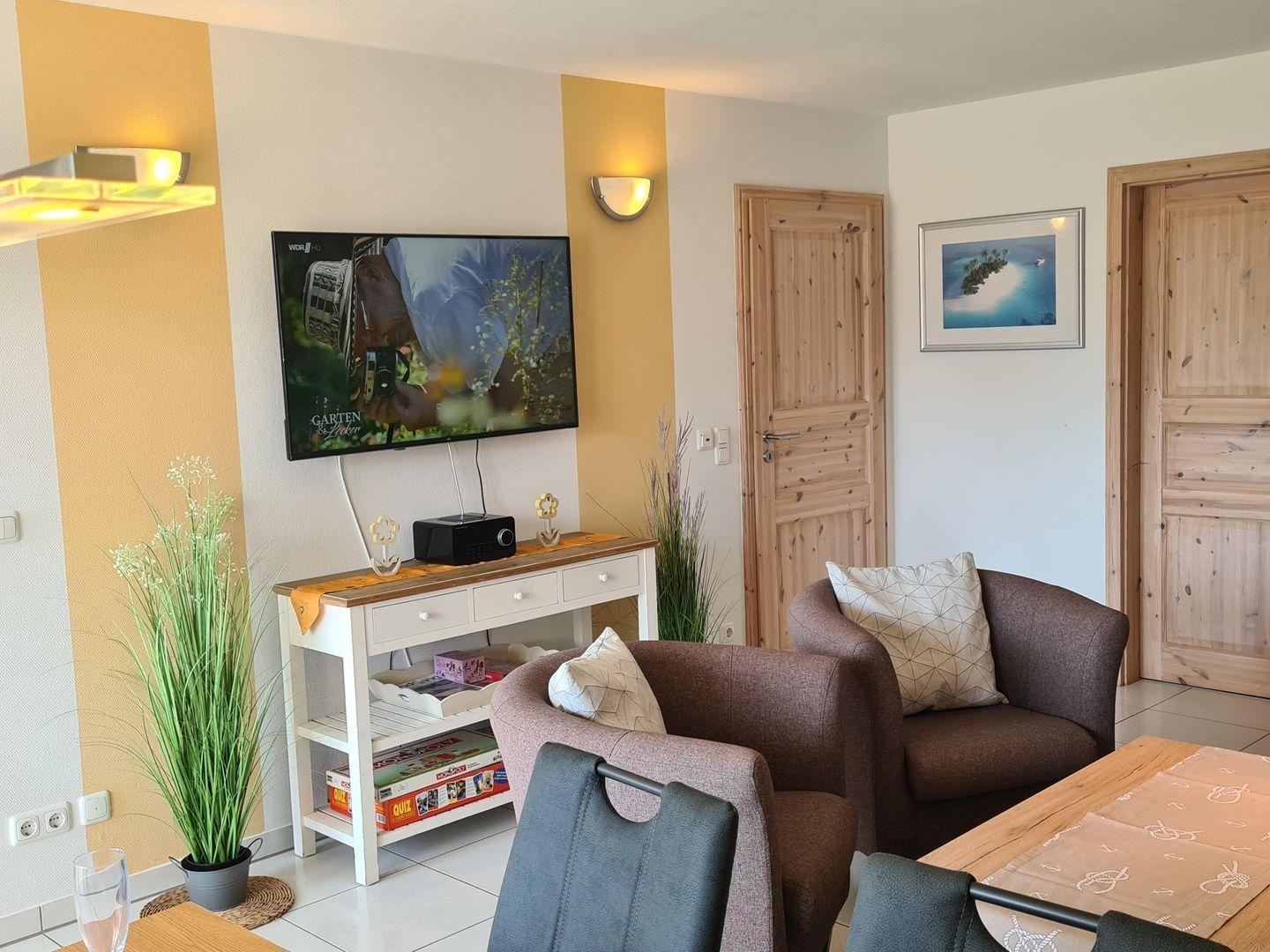 Wohnzimmer mit Fernseher, Sitzgelegenheiten und Tisch. Holztüren und Wandbehang.