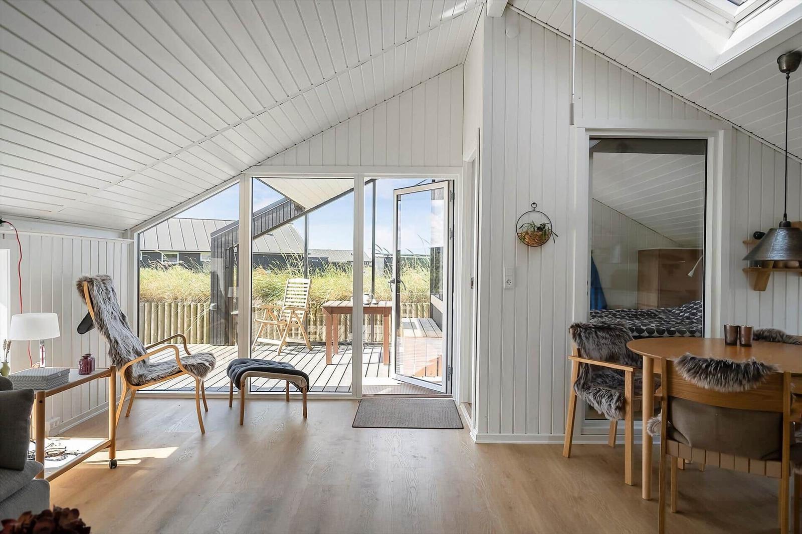 Wohnbereich mit Essbereich, großes Fenster und Terrasse. Holzboden und weiße Wände.
